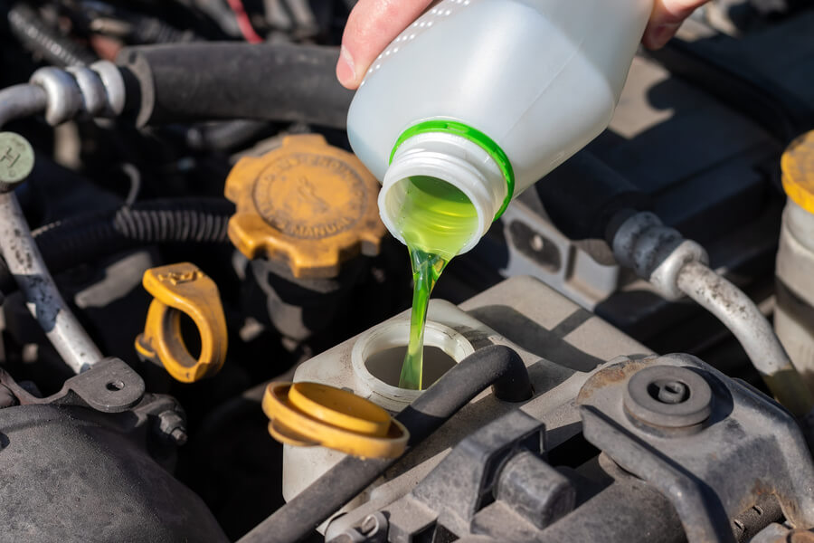 A zoomed-in photo of an engine with a hand pouring green liquid from a light gray container into an engine reservoir. The liquid is engine coolant.