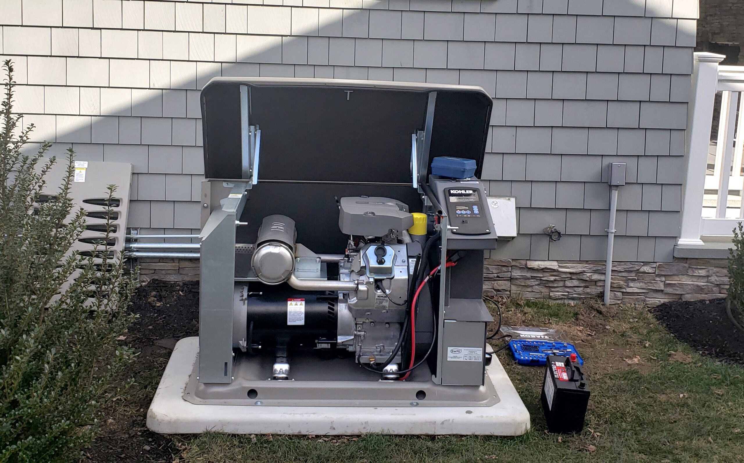 A photo of an opened kohler residential generator with a set of tools and testing equipment laying on the grass to the side of it. The side wall and foundation of a home are visible in the background.